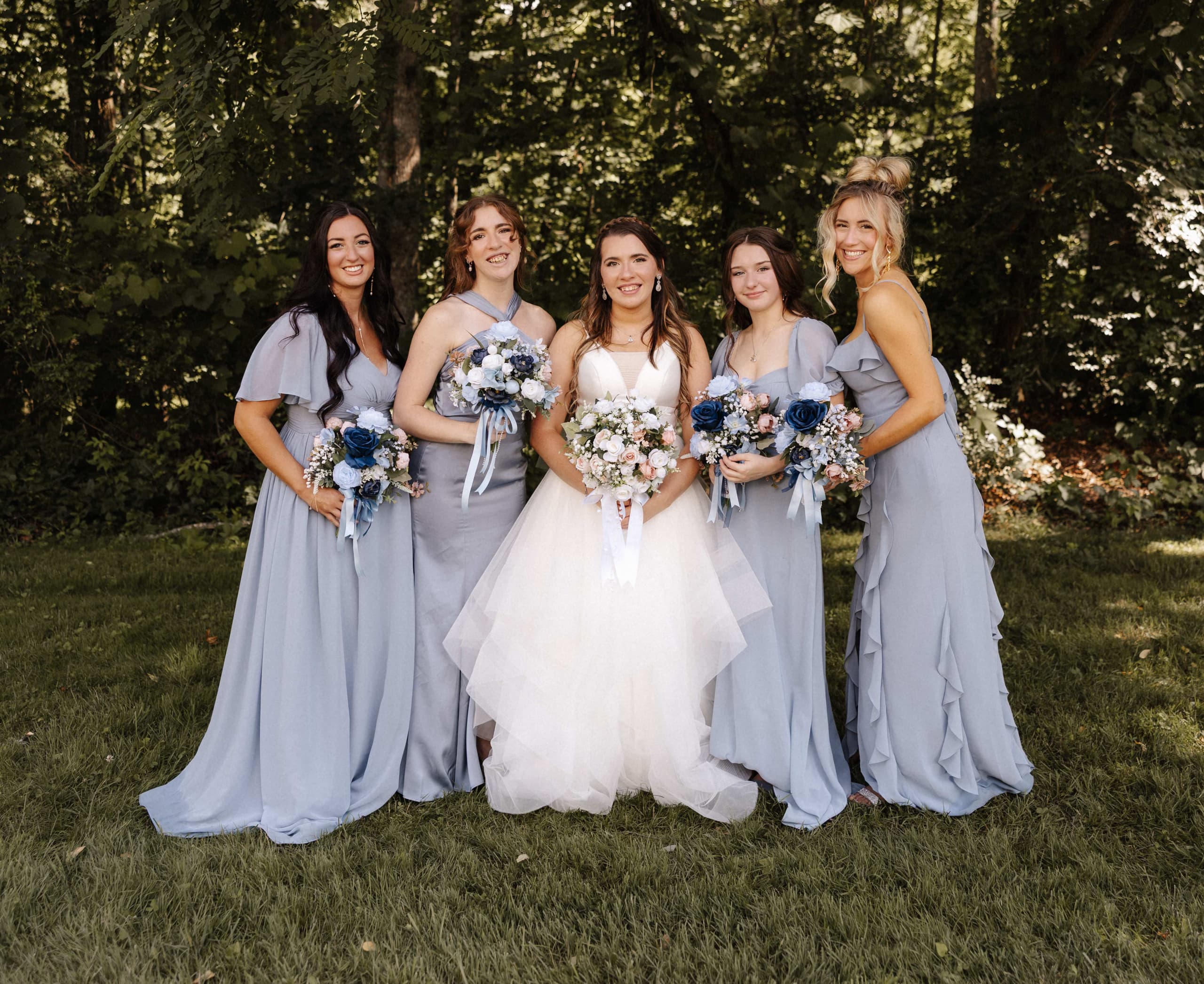 Bride with four bridesmaids in dusty blue dresses holding bouquets in a wooded setting
