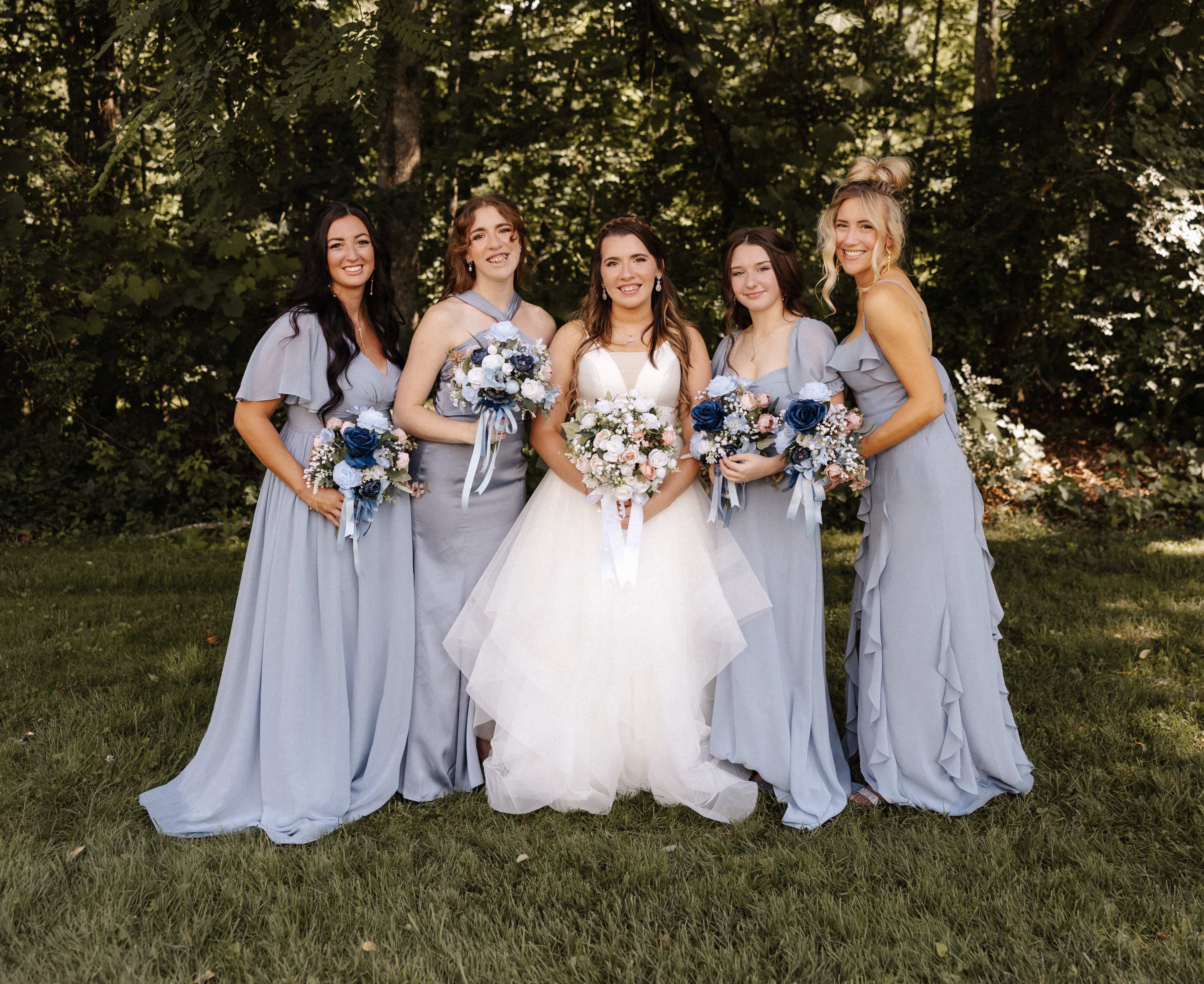 Bride with four bridesmaids in dusty blue dresses holding bouquets in a wooded setting