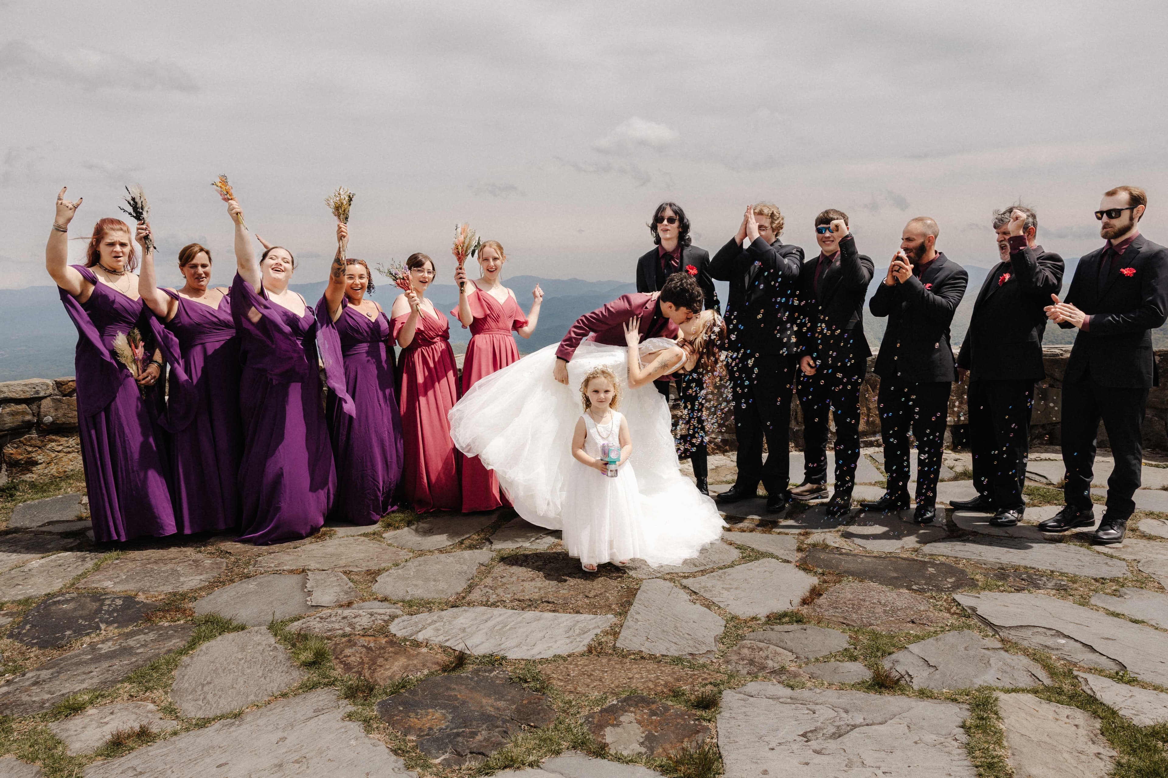 Bride and groom sharing a dip kiss surrounded by their cheering wedding party on a mountaintop overlook