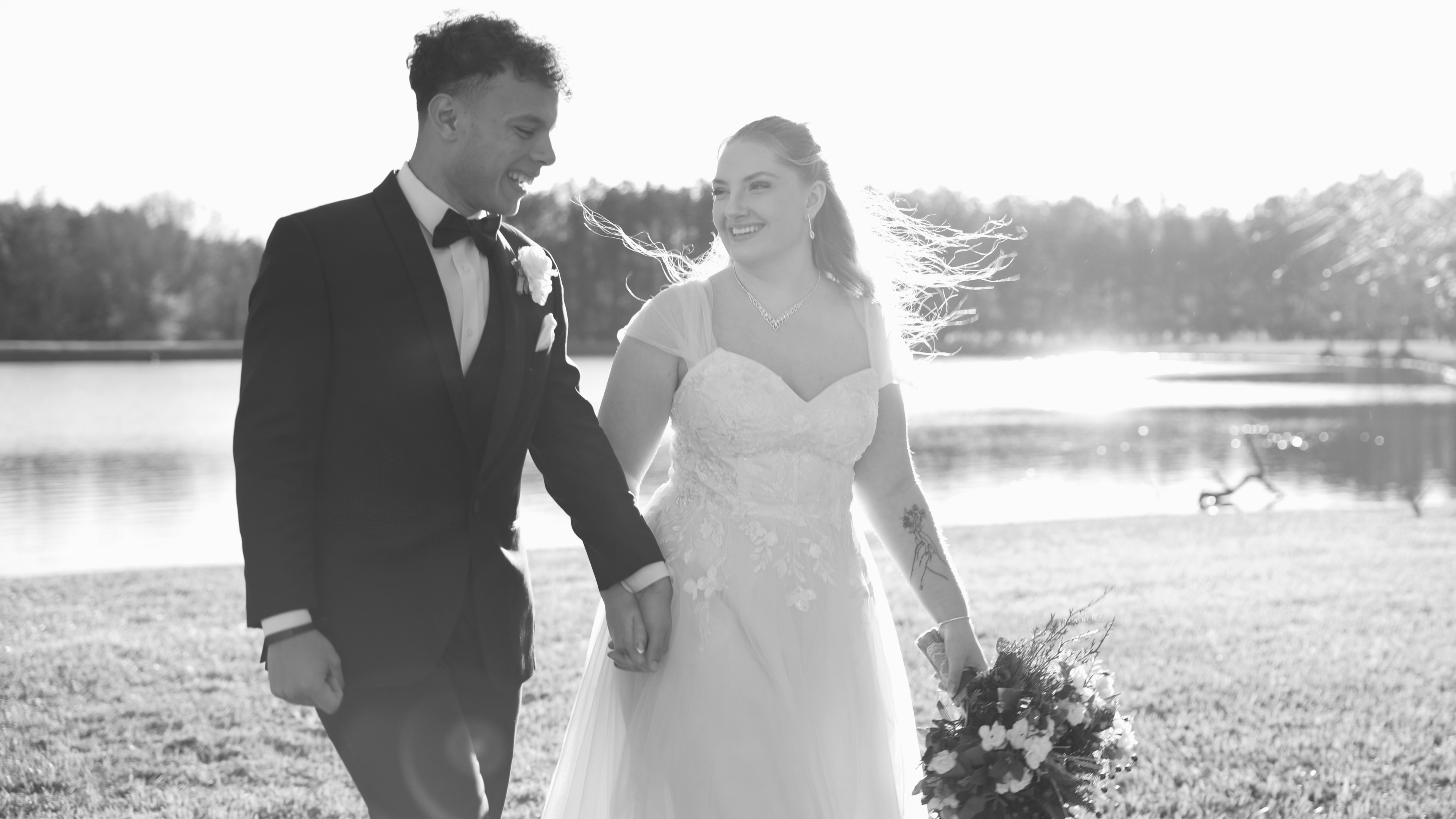 Black and white photo of bride and groom walking together by a lake at Sunset Farms